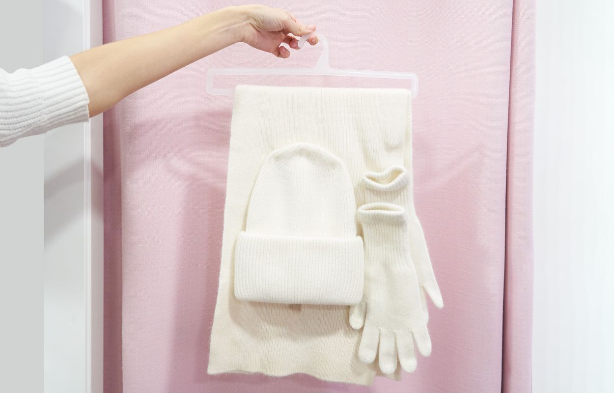 Conjunto de invierno en color crema compuesto por bufanda, gorro y guantes, sostenido sobre una percha.